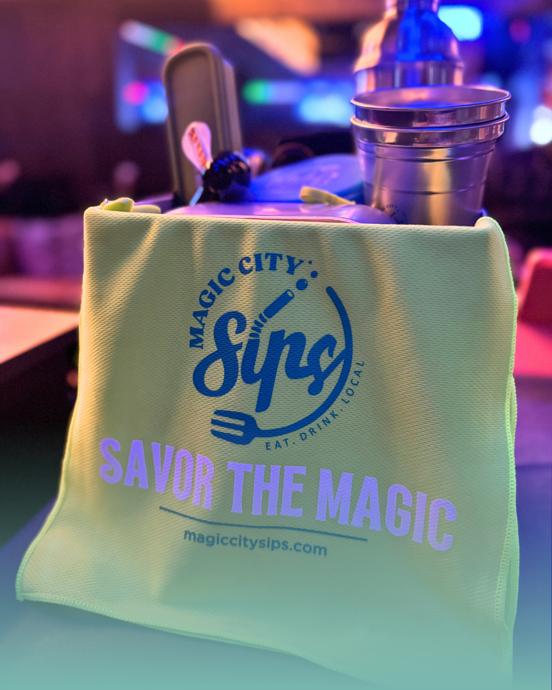 A bright neon green promotional bag displaying the Magic City Sips logo and the text 'SAVOR THE MAGIC' with the website URL, sitting next to a stainless steel cocktail shaker and cups.